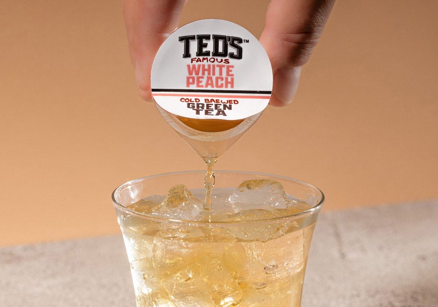 Person pouring a Ted's famous white peach green tea pod into a glass of ice
